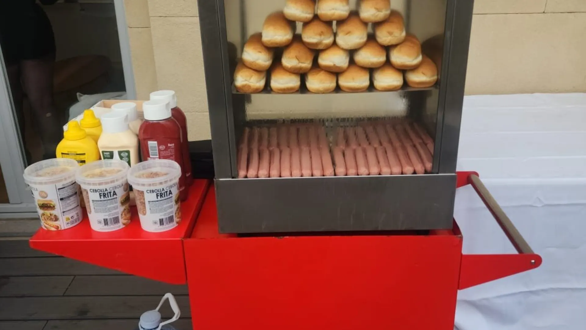 Carrito de hot dog rojo con panecillos y salchichas en la vitrina y salsas a un lado