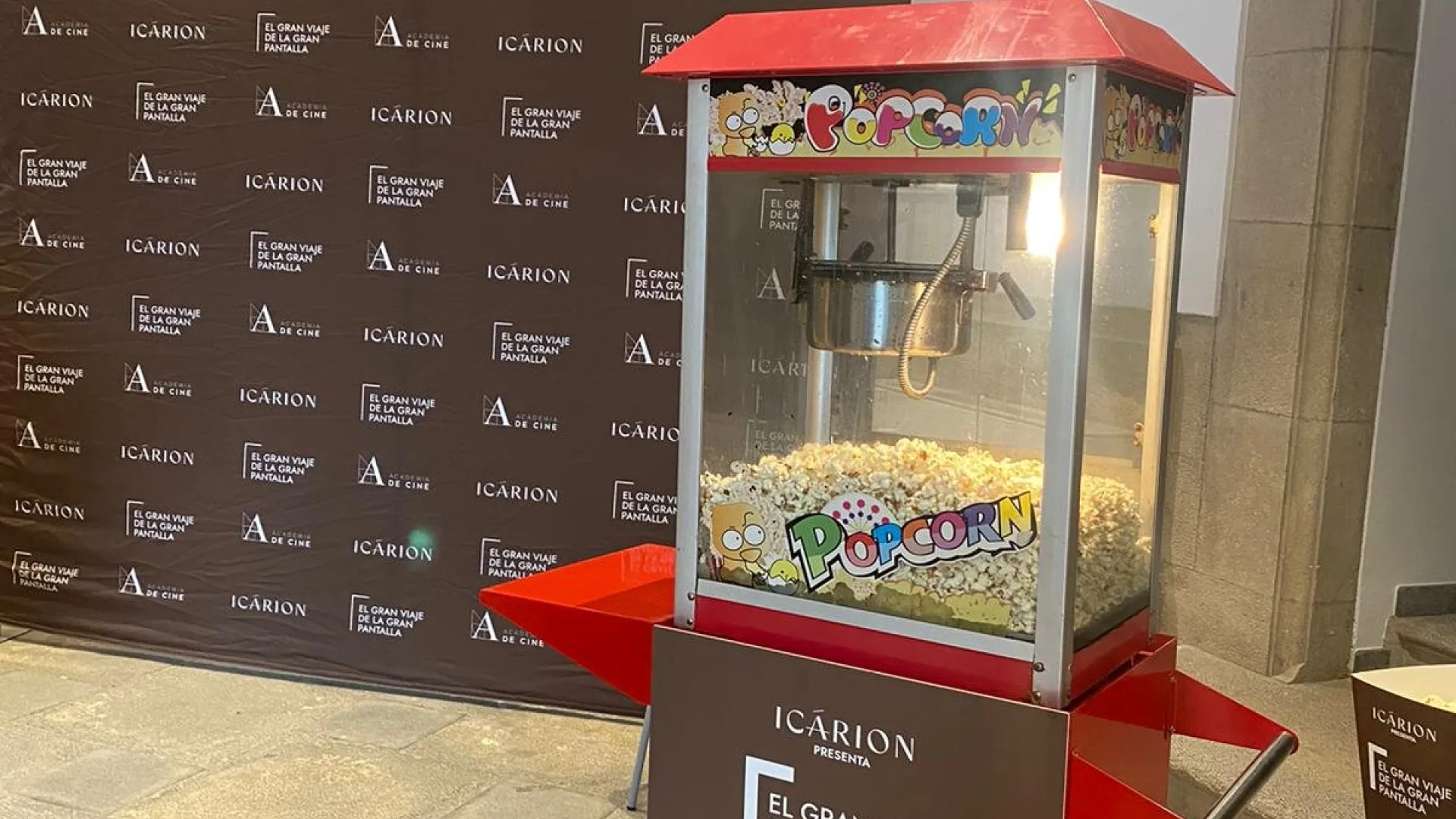 Carrito de palomitas rojo lleno de palomitas, con panel personalizado, en la entrada de un evento de cine