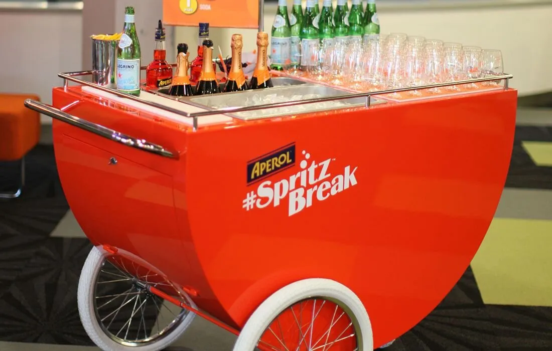 Carrito de spritz rojo con botellas y copas en un evento de interior