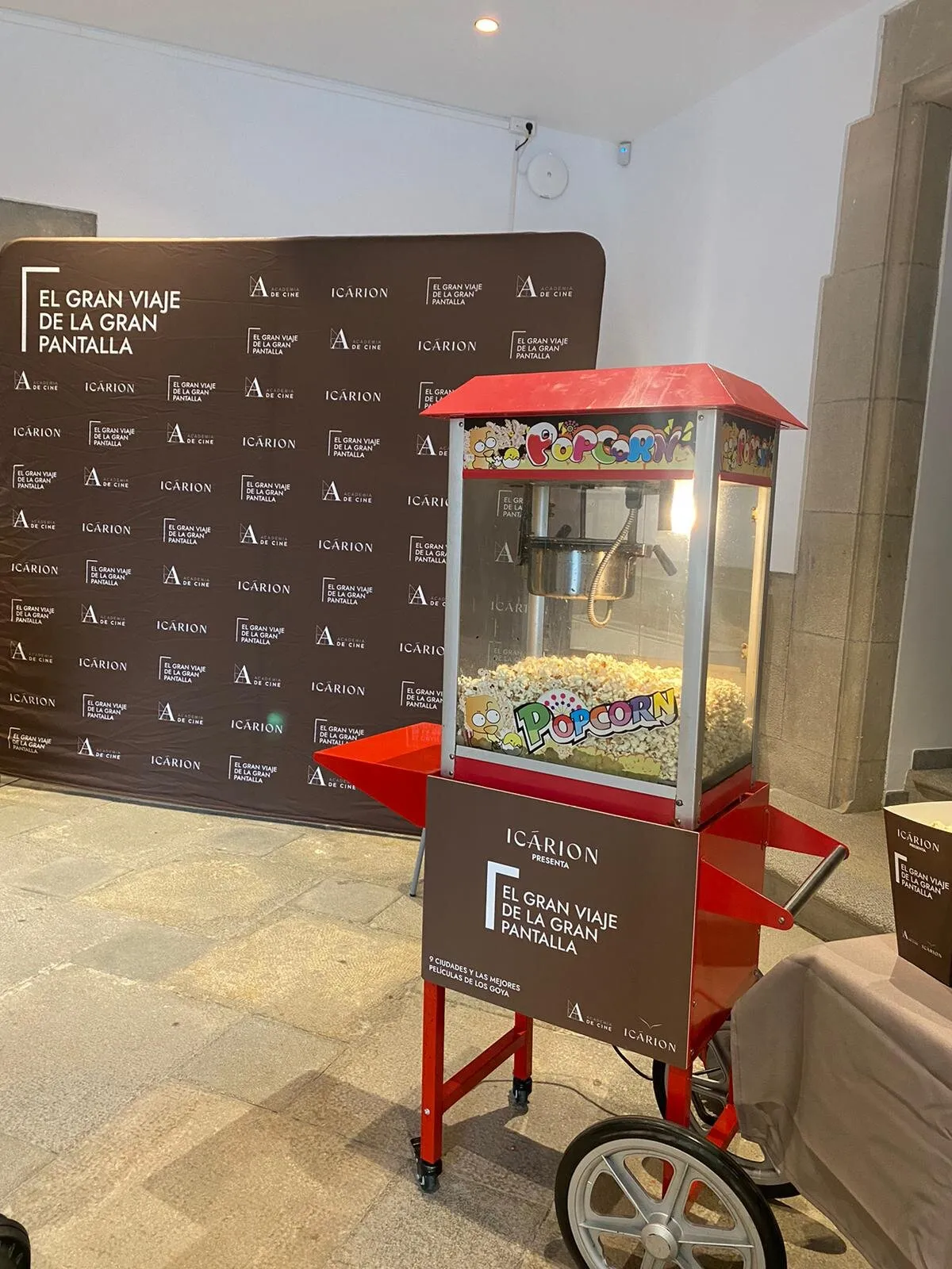 Carrito de palomitas rojo lleno de palomitas, con panel personalizado, en la entrada de un evento de cine